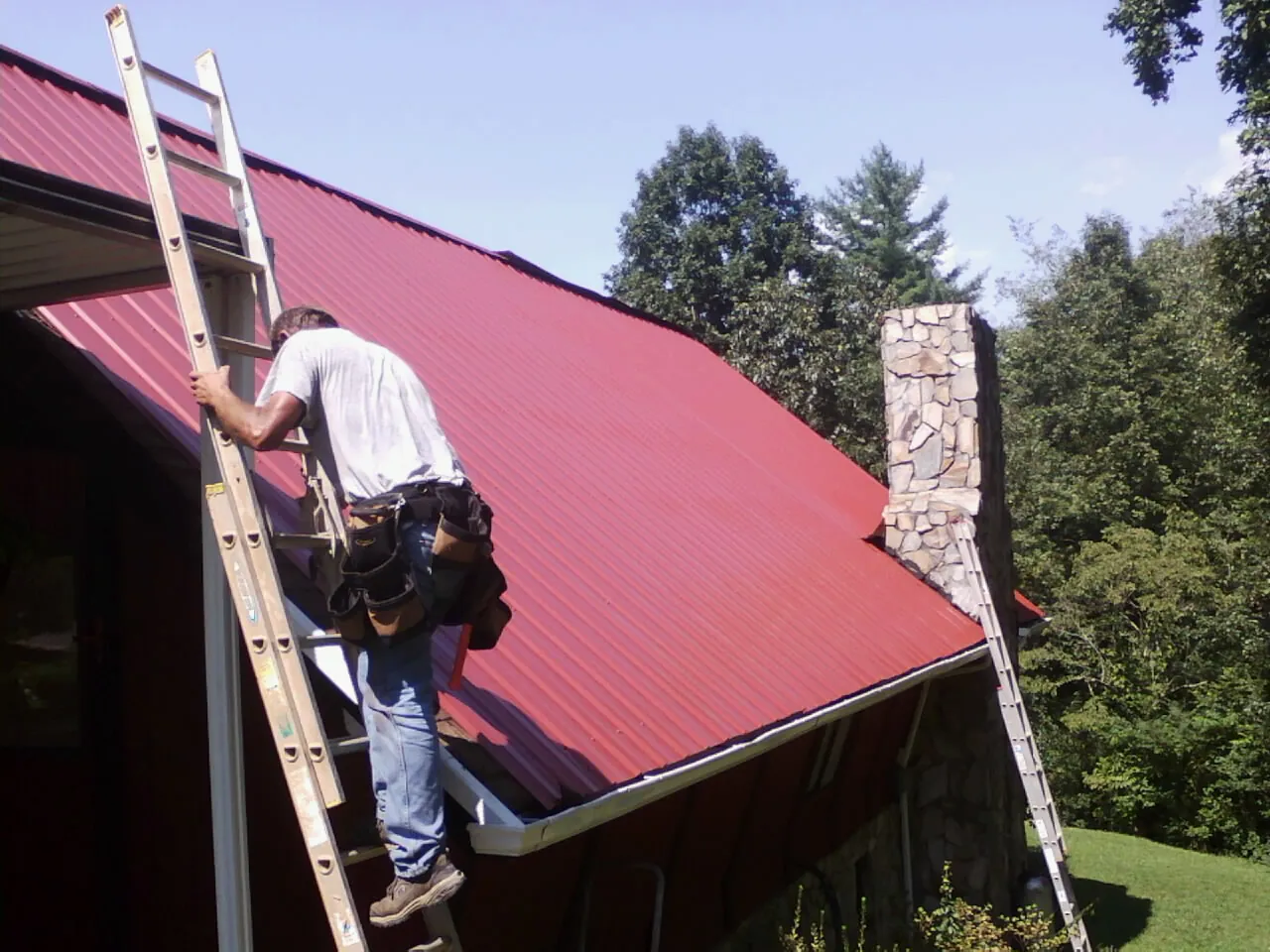 Roofer on a ladder beside a red metal roof and stone chimney — Soffit & Fascia Repair by Roofing Brothers in Harwich Port